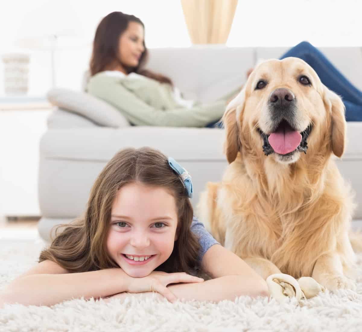 Jeune fille allongée sur un tapis avec un golden retriever, en arrière-plan une femme détendue sur un canapé - environnement familial propre et soigné par Nettoyage Impérial